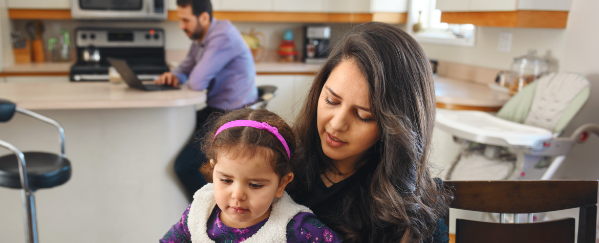 Woman sitting at kitchen table with young female child on her lap. Man sitting in background looking at laptop screen