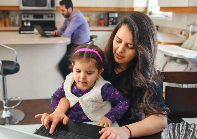 Woman sitting at kitchen table with young female child on her lap. Man sitting in background looking at laptop screen