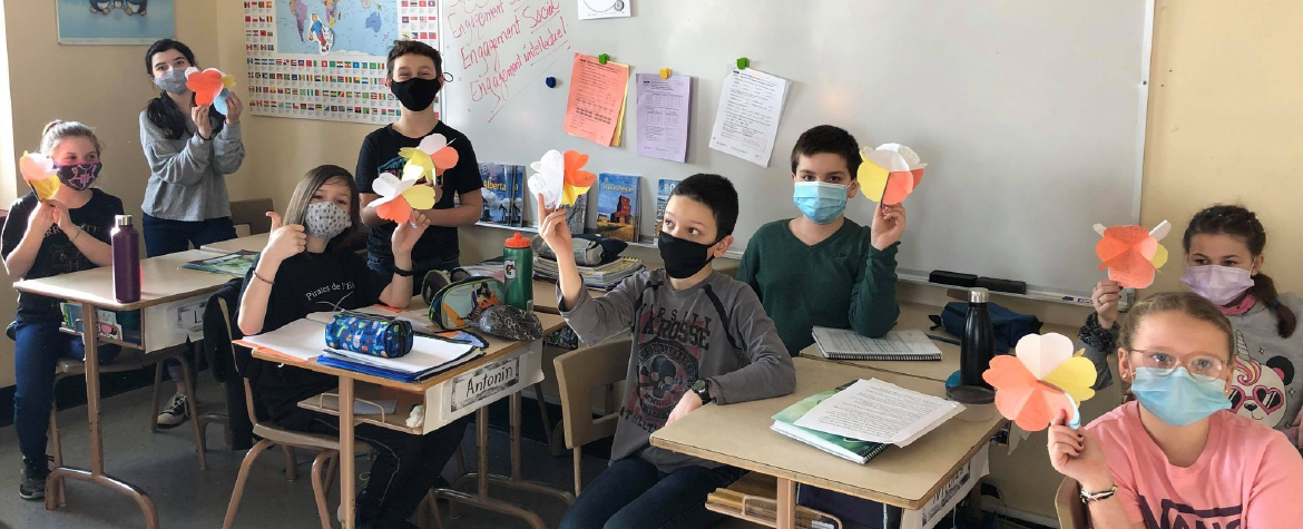 Young masked children sitting at classroom desks holding up artwork